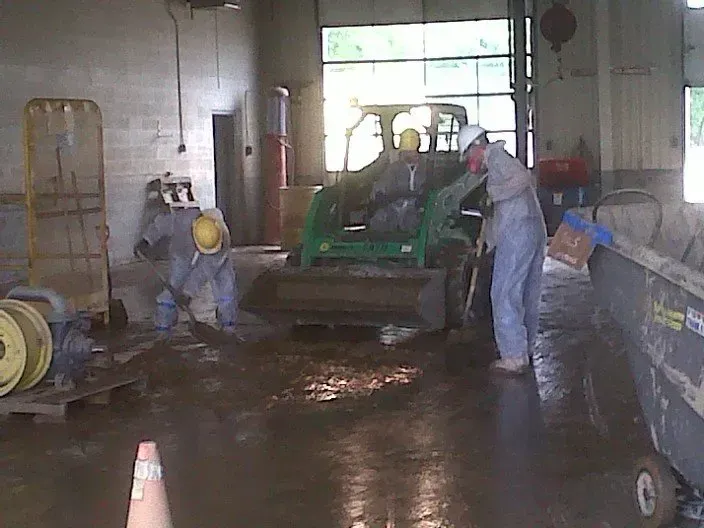 Workers in protective suits and hard hats cleaning a wet industrial area with a skid steer loader.