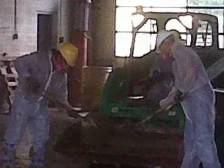 Two workers in protective suits and masks shoveling near a green machine in a factory.