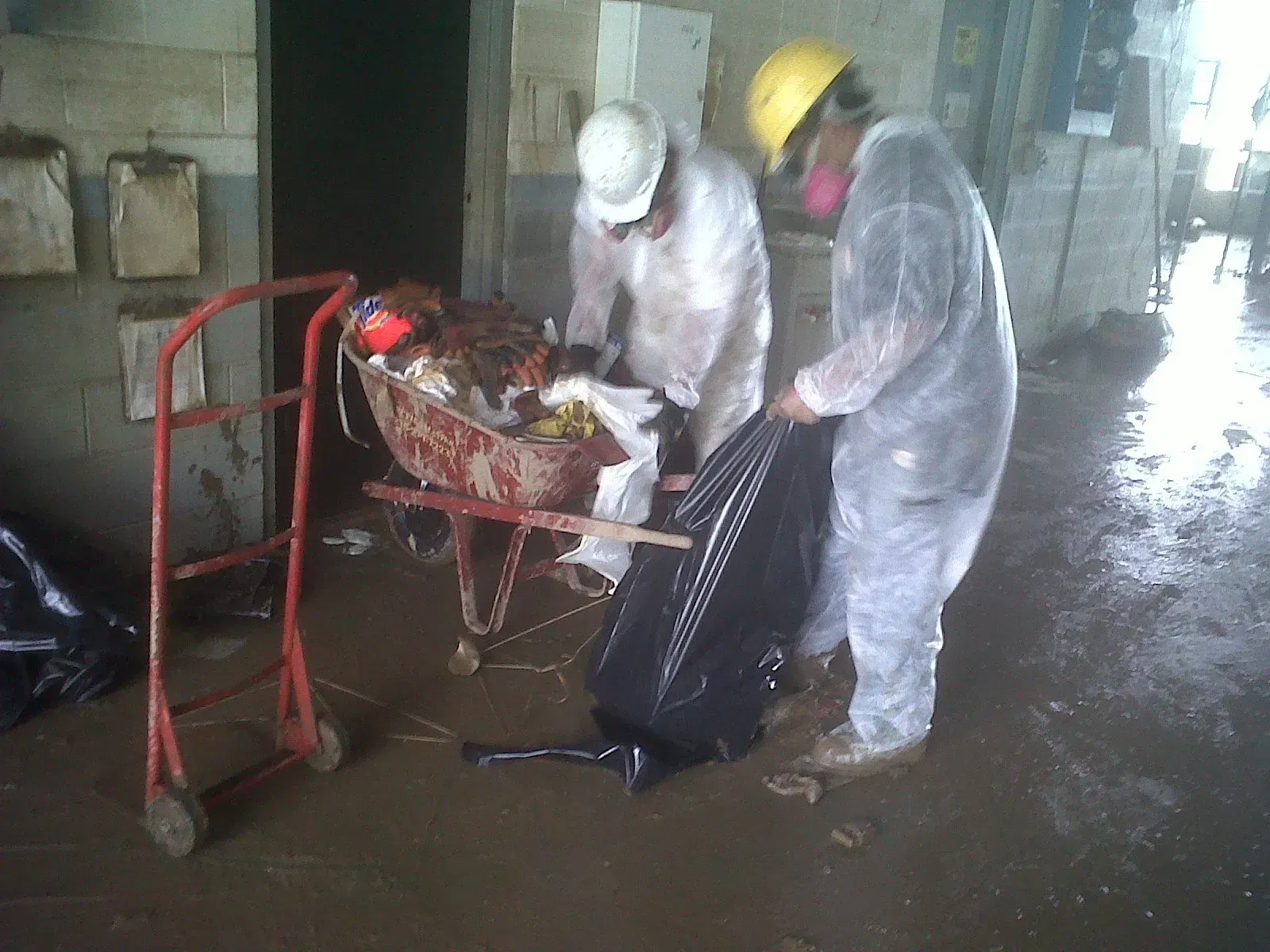 Two workers in protective suits loading debris into a black bag, using a wheelbarrow in a flooded area.