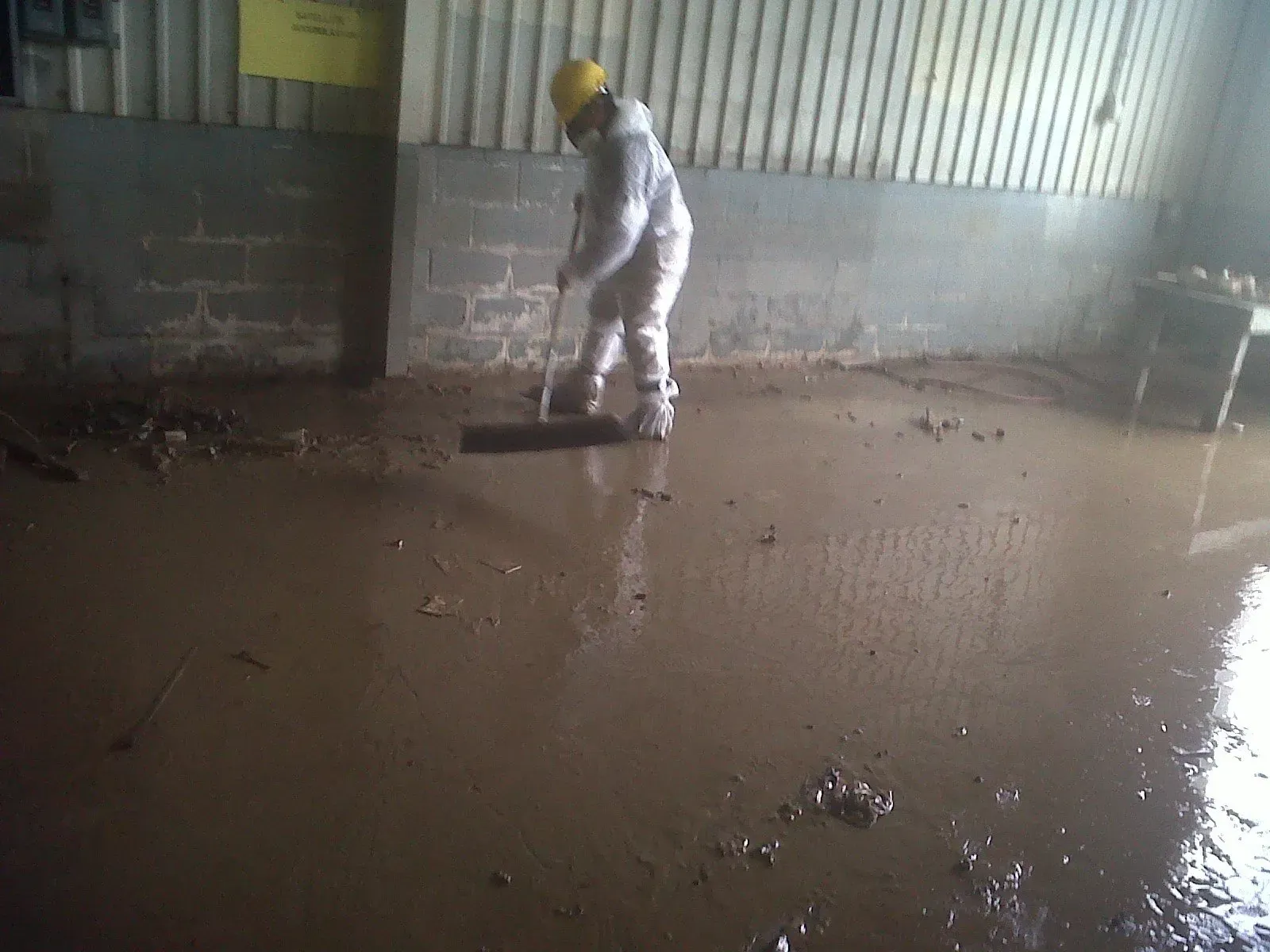 Person in protective suit sweeping a muddy, flooded industrial area.