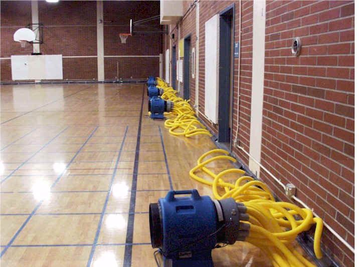 Row of blue air movers with yellow hoses drying a gymnasium's wood floor against a brick wall.