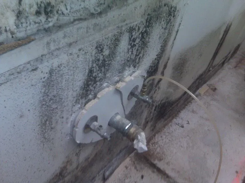 Close-up of a damaged wall with a metal pipe and mounting plate. Dark stains and possible mold present.