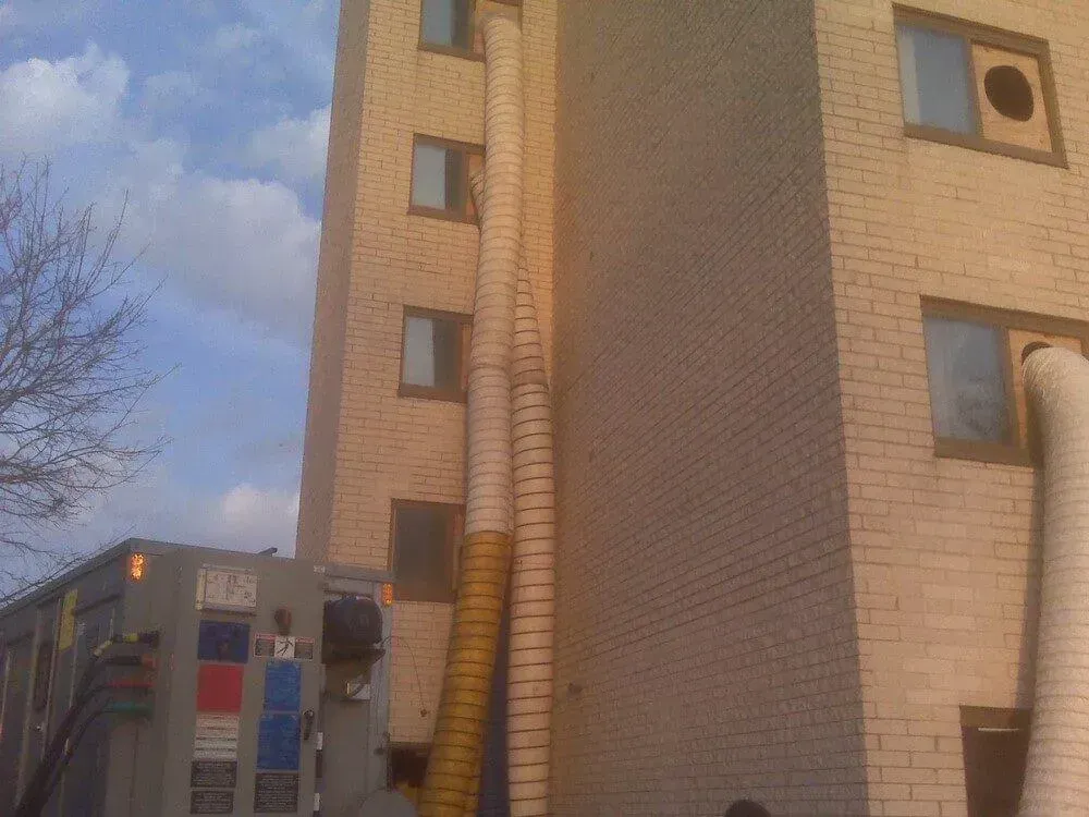Building with air duct hoses extending to windows, attached to a large truck on a sunny day.
