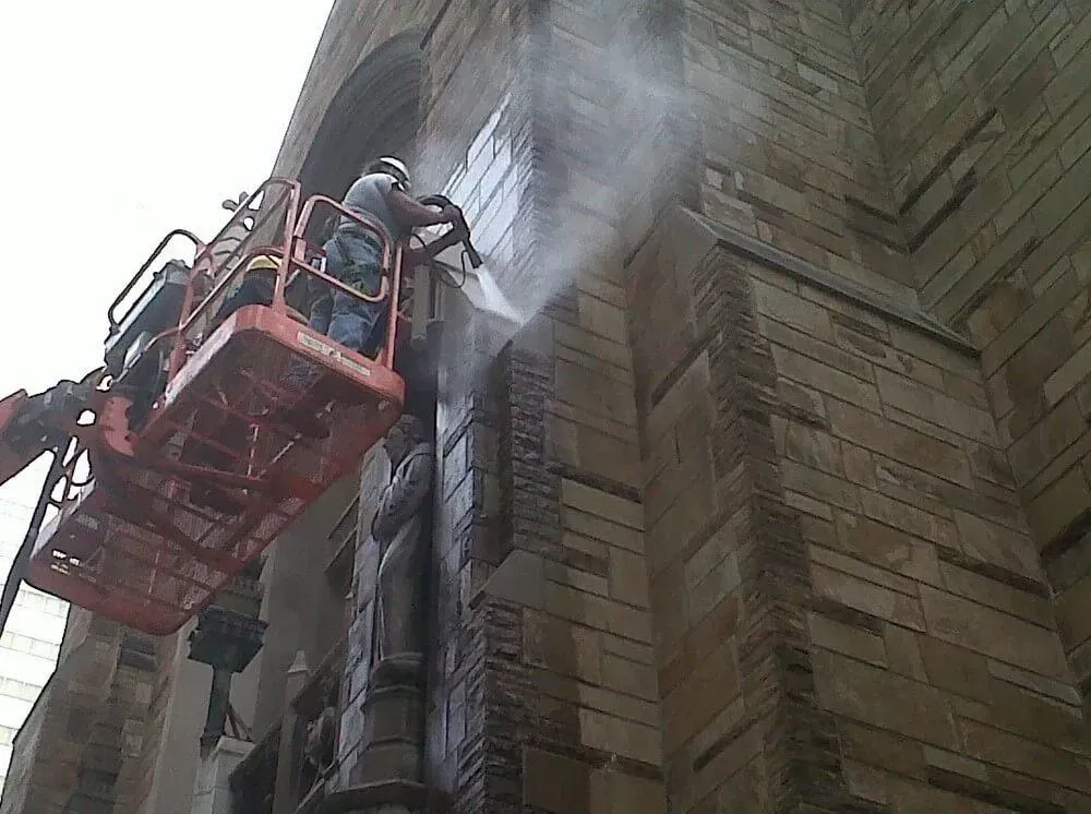 Worker in a lift washing a stone building facade with a pressure washer.