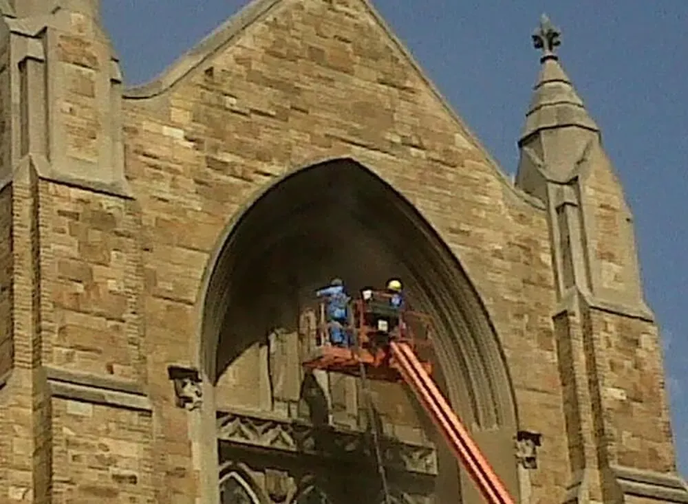 Workers on a lift repairing the arched facade of a stone church.