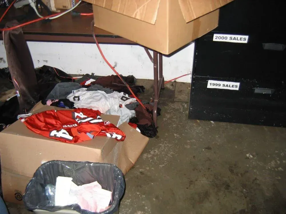 Box and trash can in a cluttered storage space with wet floor. Papers, clothes, and a cabinet labeled 