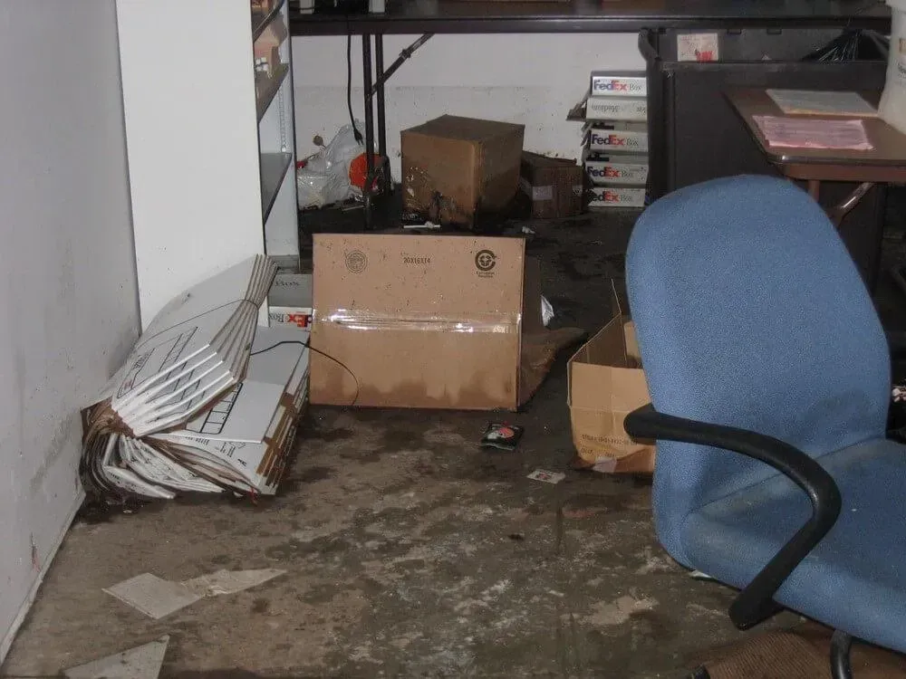 A cluttered room with damaged boxes, chair, and a dirty floor, possibly flooded.