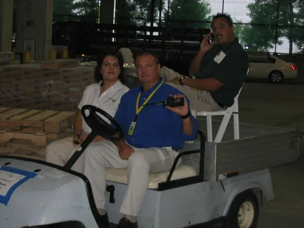 Three people sit in a utility cart. One holds a video camera, another is on the phone. Setting is outdoors, likely a work area.