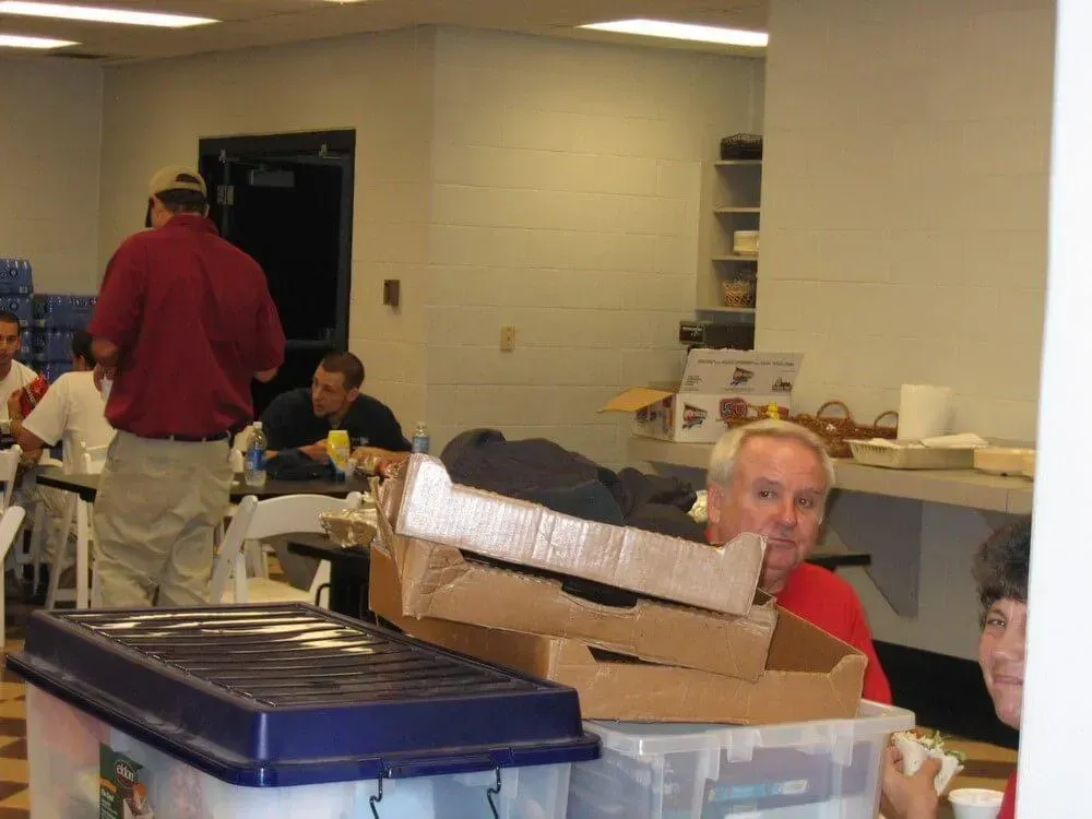 People in a room, some seated at tables. Man in red shirt smiles at camera. Boxes and bins in foreground.