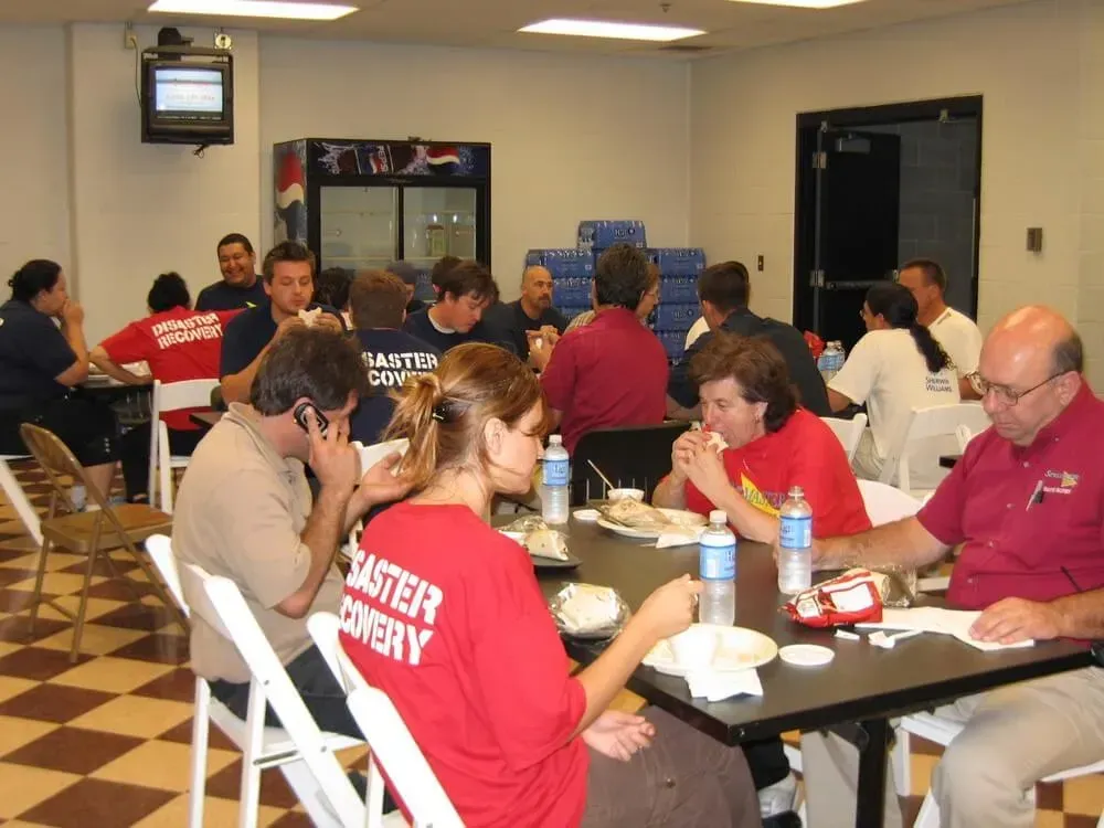 Group of people eating at tables in a room, likely a disaster recovery center.