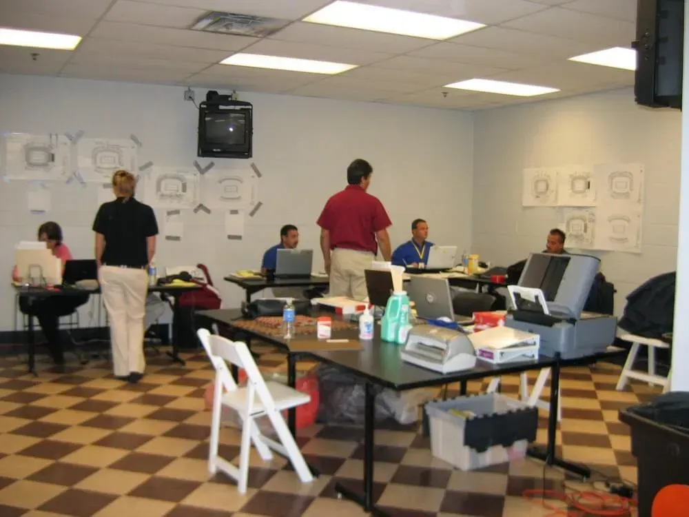 People working on laptops at tables in a room with a checkered floor. Some stand and some sit. Schematics are on the wall.