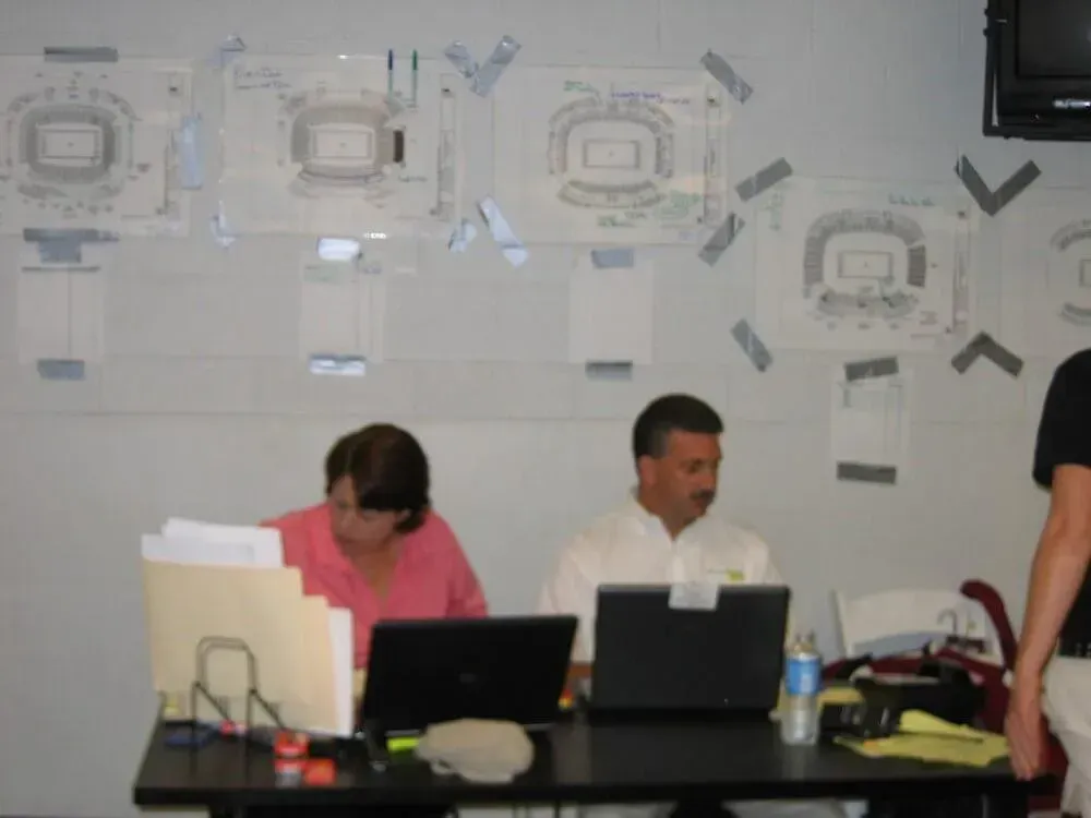 Two people at a table with laptops, blueprints on a wall. Office setting.