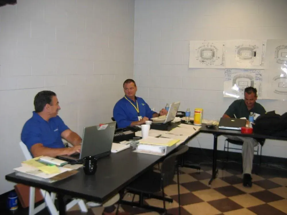 Three men at a table with laptops, working in a room with stadium diagrams on the wall.
