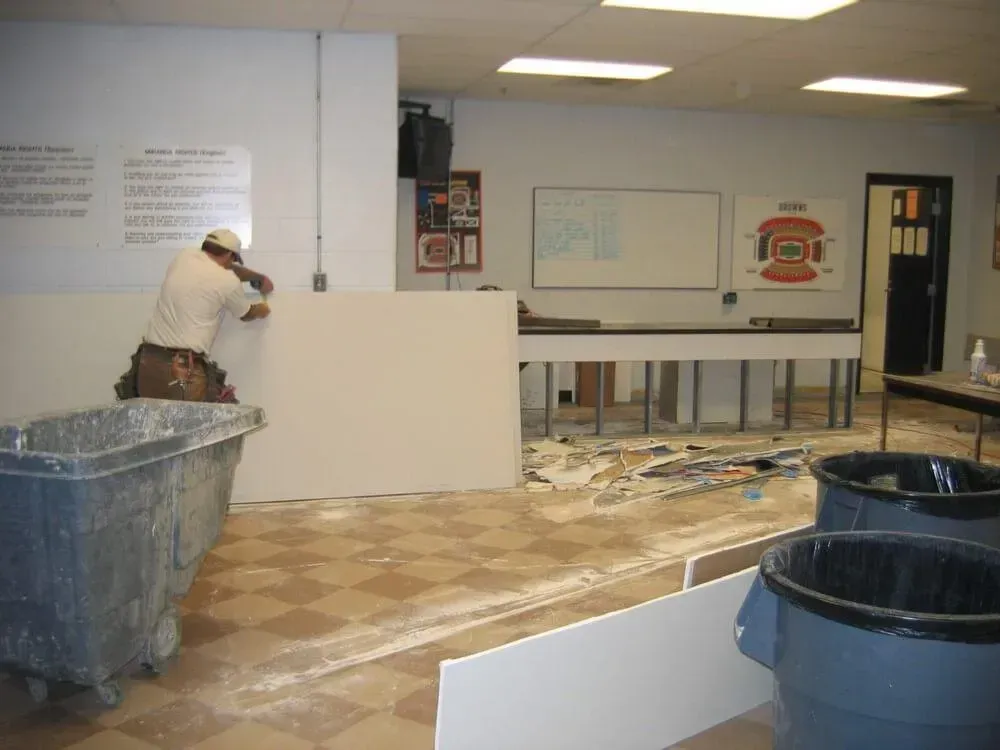 A worker installs drywall in a room with a checkered floor, trash bins, and a whiteboard.