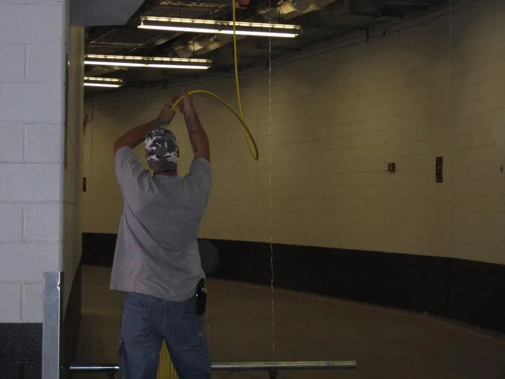 Man in a tunnel holding a yellow cord overhead. Setting is an industrial interior with beige walls.