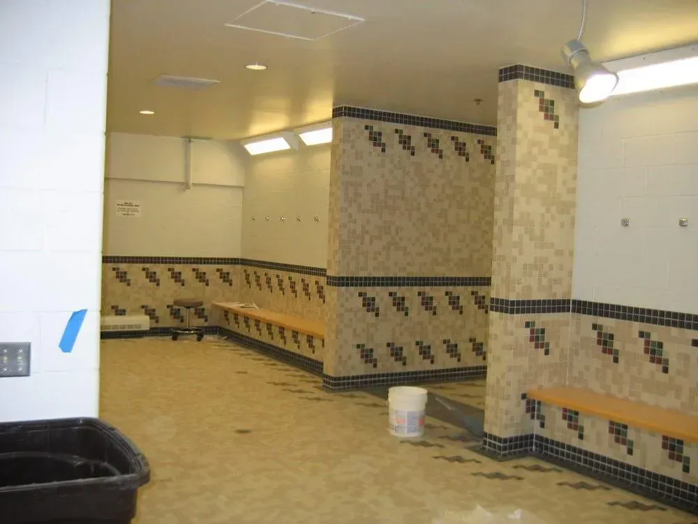 Public shower room with beige tile walls, benches, and a black bin.