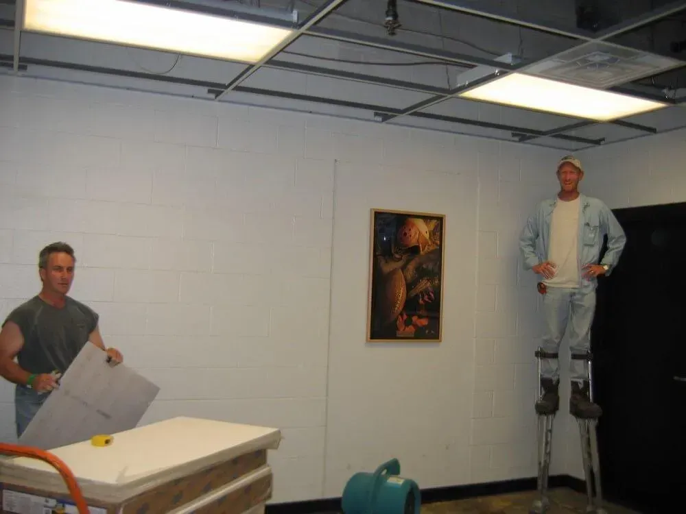 Two men in a room; one on stilts, one holding a large sheet of material. A painting hangs on the wall.