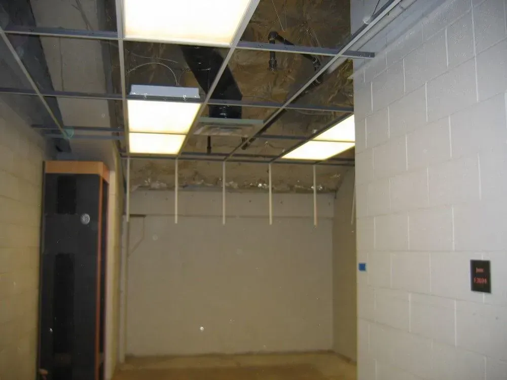 Empty room with dropped ceiling and fluorescent lights, light gray walls, and dark cabinet.