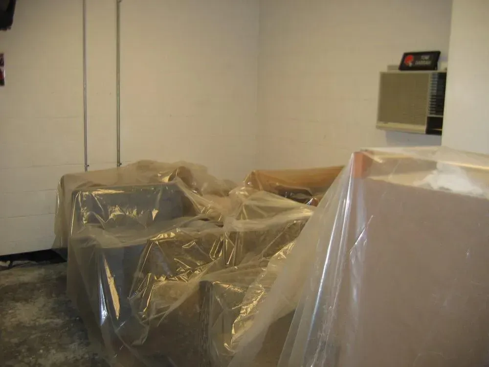 Furniture covered in plastic sheeting in a room with white walls. An air conditioning unit is on the wall.