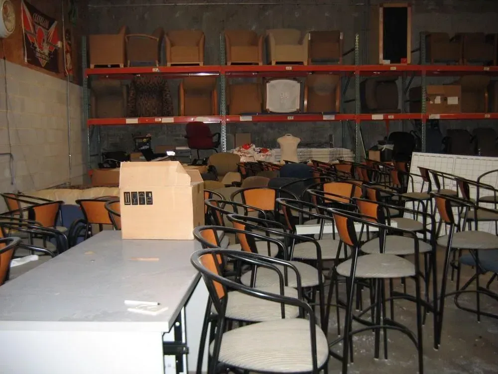 Warehouse interior with chairs, storage racks filled with furniture, and a large white desk.