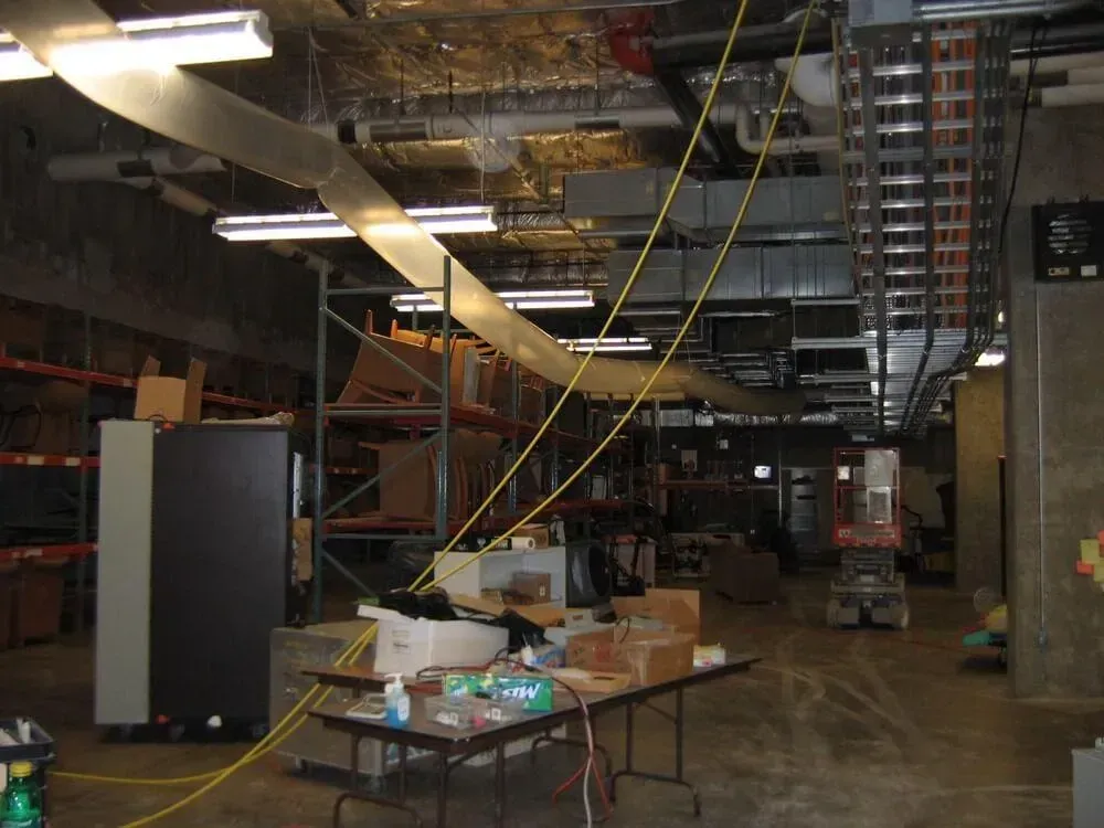 Warehouse interior with exposed pipes, electrical wiring, and a table with supplies.