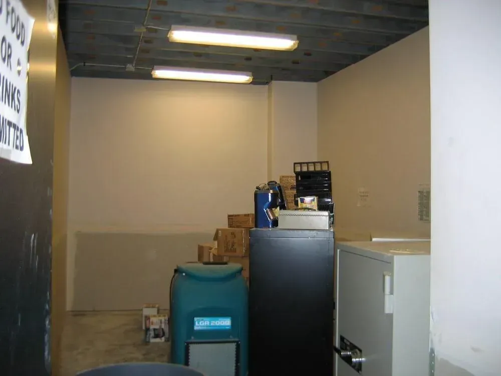 A small room with beige walls, fluorescent lights, and various equipment like a safe and trash bins.
