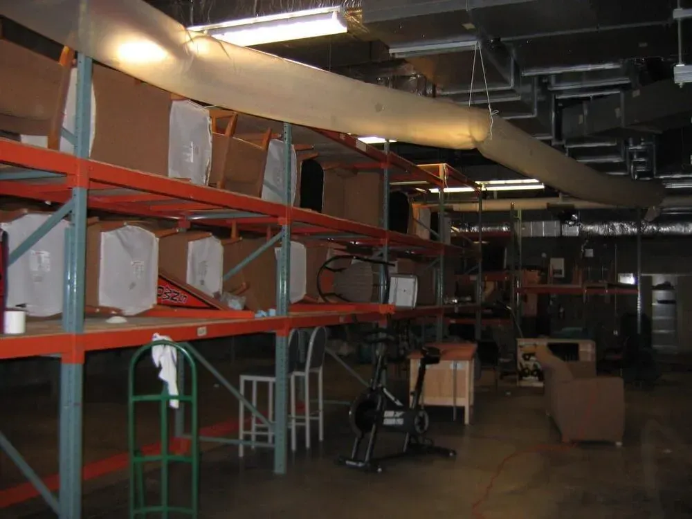 Warehouse with shelving, boxes, and a fabric ventilation duct. Exercise bike and furniture are visible.