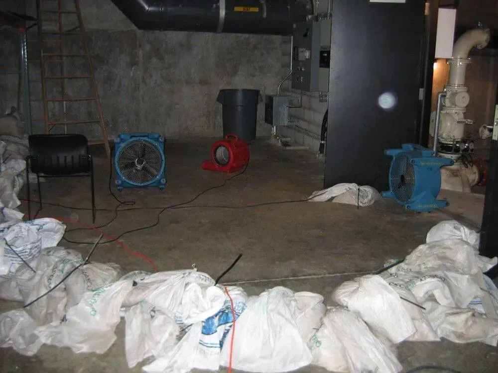 Basement with three fans and debris bags on floor, possibly related to water damage or cleanup.