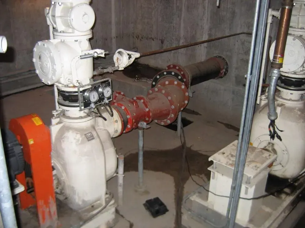 Industrial pump system in a concrete room with pipes, gauges, and wet floor.