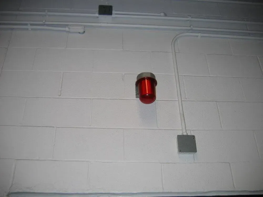 Red fire alarm light mounted on a white cinder block wall. Electrical conduit visible.