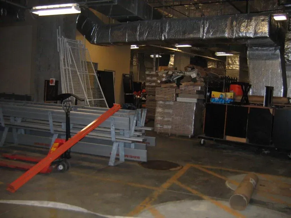 Warehouse with metal beams, stacked boxes, and equipment. Ductwork and stairs visible.