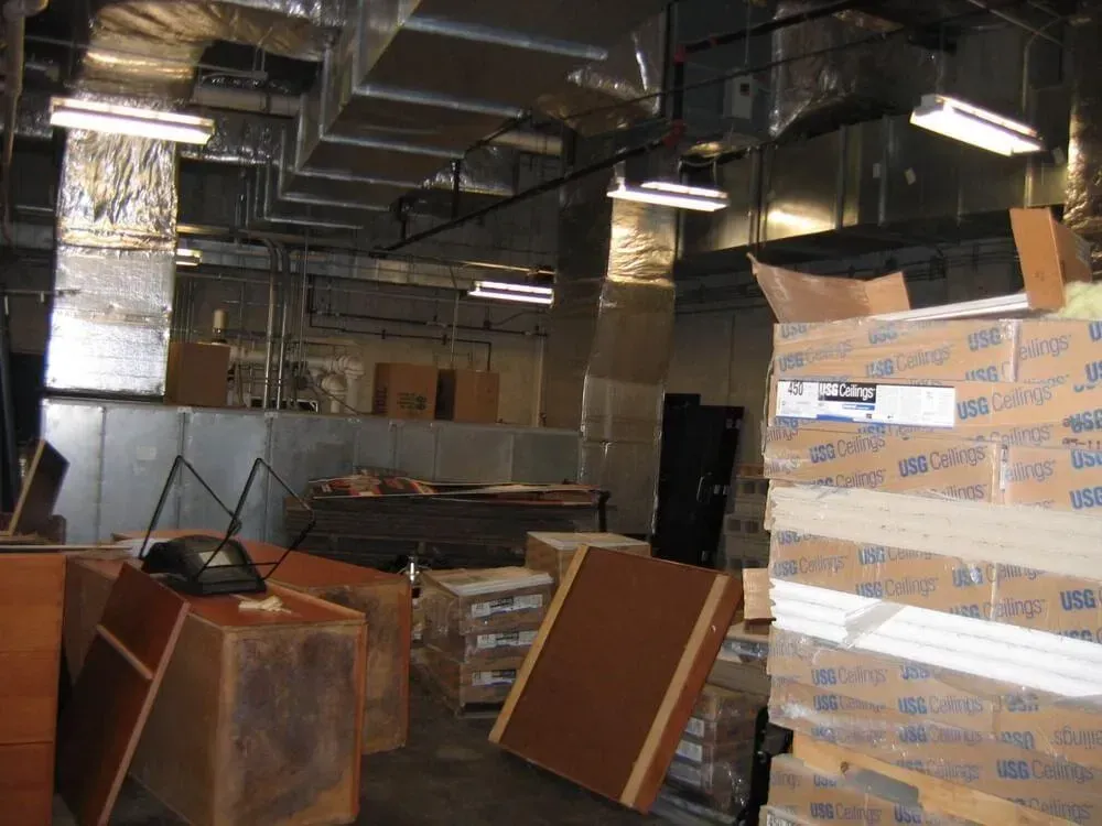 Basement storage area filled with boxes, wooden furniture, and ductwork. Fluorescent lights illuminate the space.