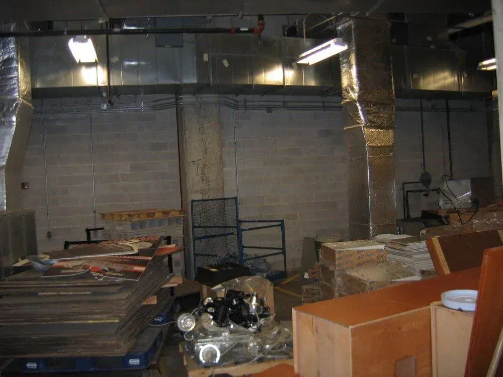 Cluttered storage area with cinder block walls, exposed ducts, and various boxes and materials.