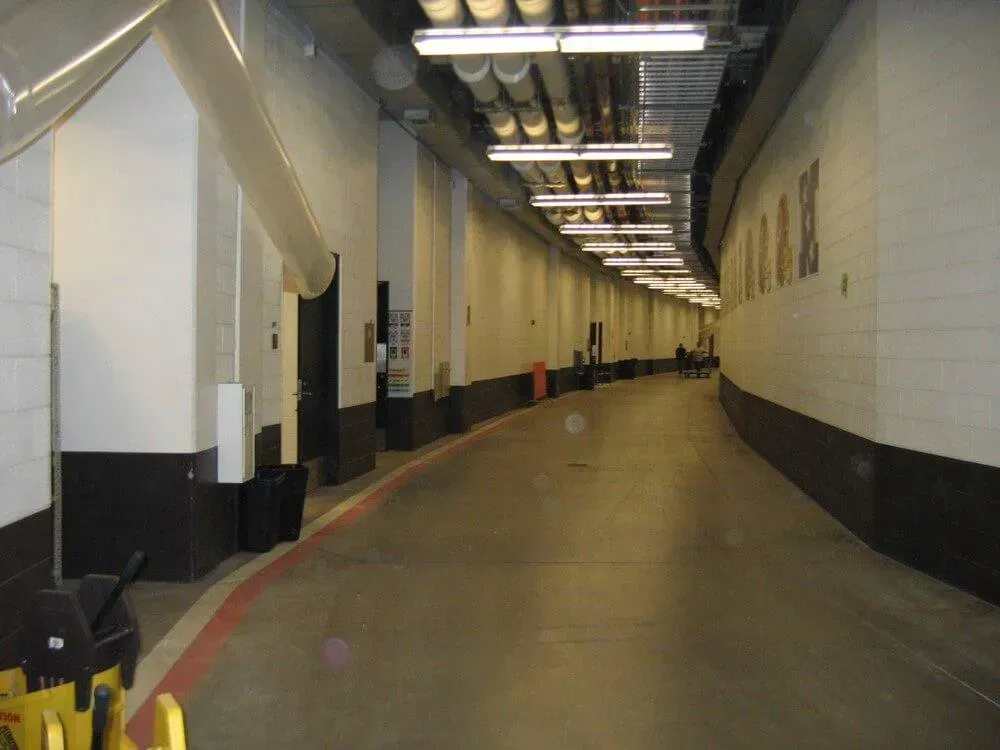 Long, curving, concrete hallway with white walls and overhead lighting. Dark brown baseboards and a red line.