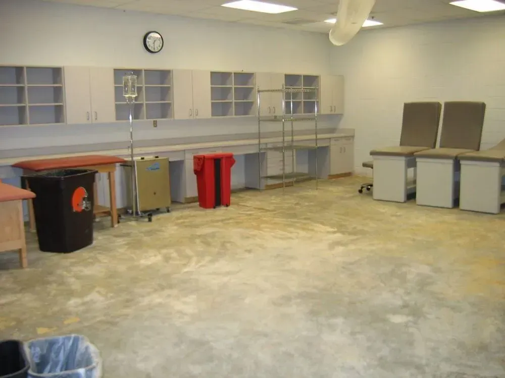 Empty medical exam room with cabinets, counters, and equipment on a concrete floor.