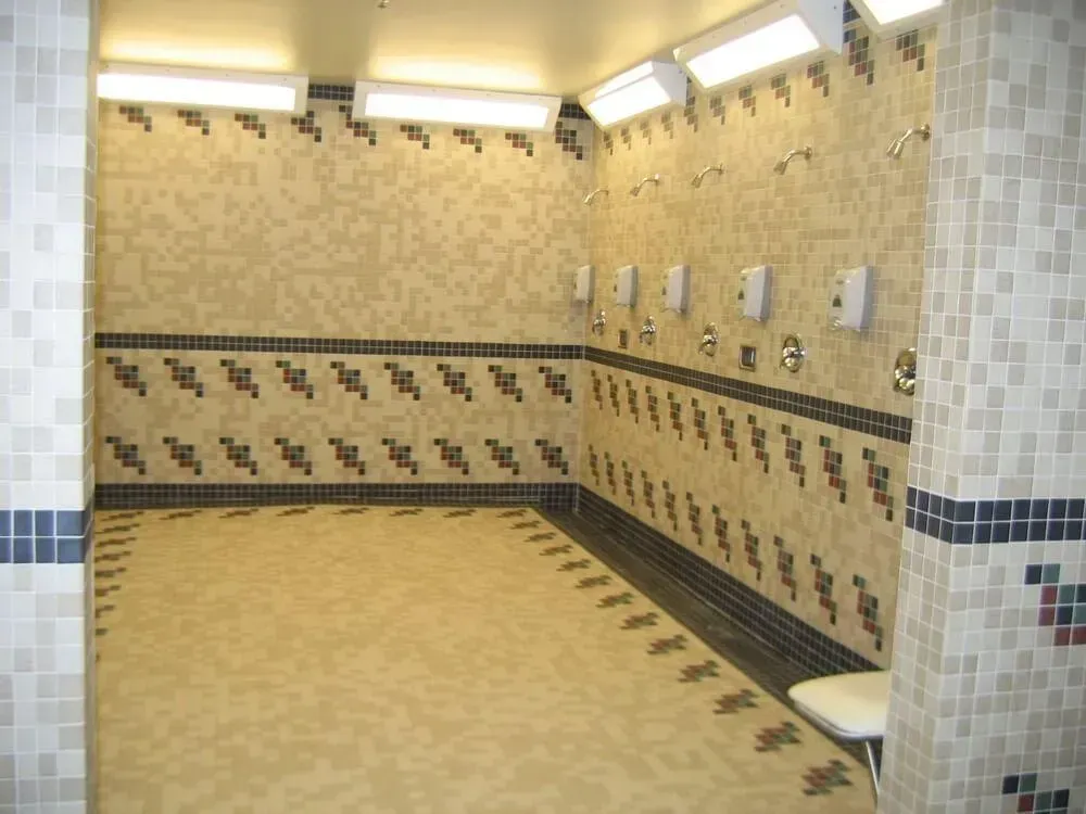 Public shower room with beige and blue tiled walls and floor, multiple shower heads, and overhead lights.