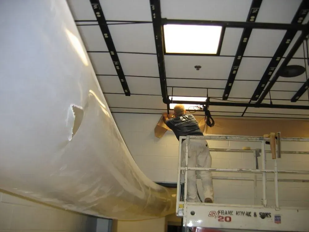 Person on a lift installs white material to a wall, working under ceiling lights.