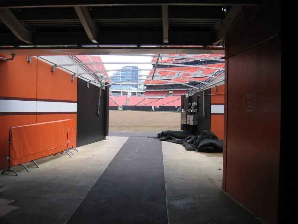 View from an interior corridor leading toward a baseball stadium field. Orange walls, black flooring, and stadium seating are visible.