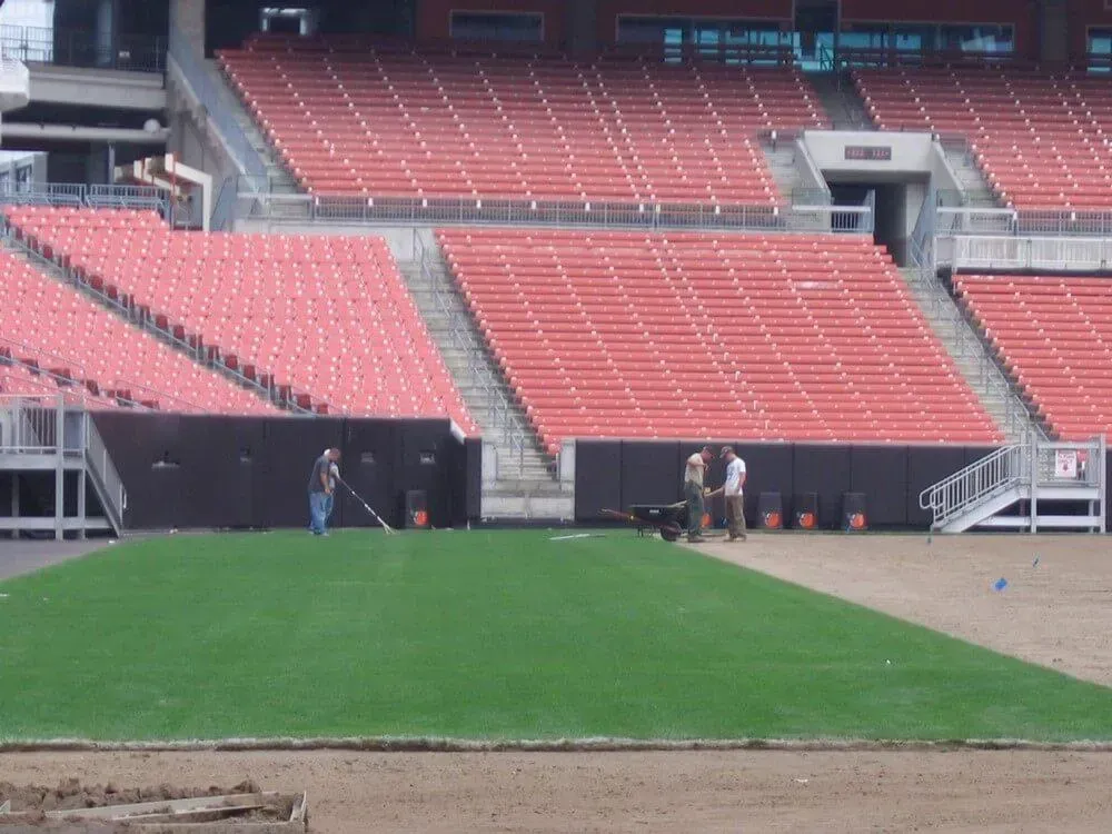 People working on a green turf area in a stadium with red seating.