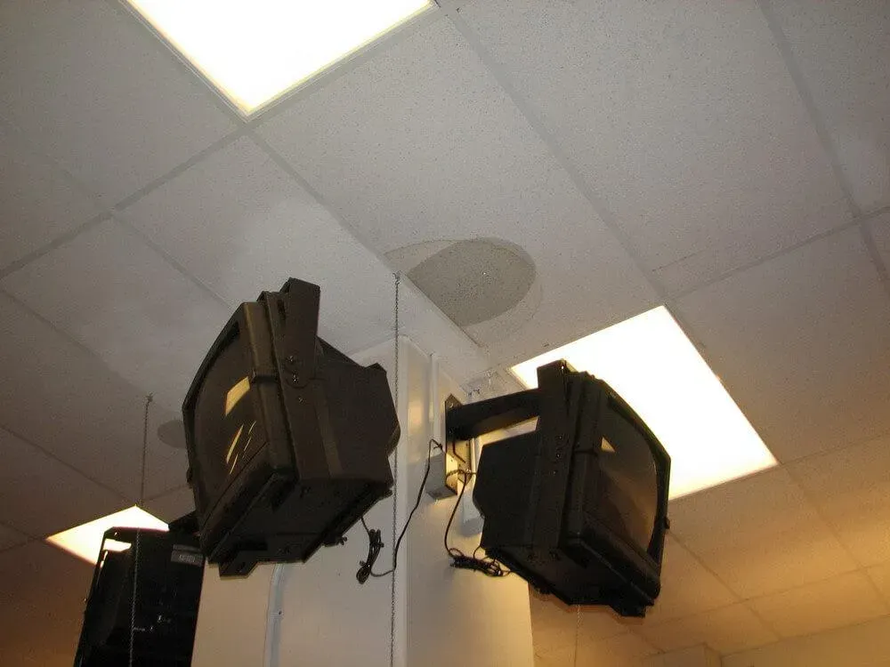 Several old, boxy TVs mounted on a white wall and ceiling with square fluorescent lights.