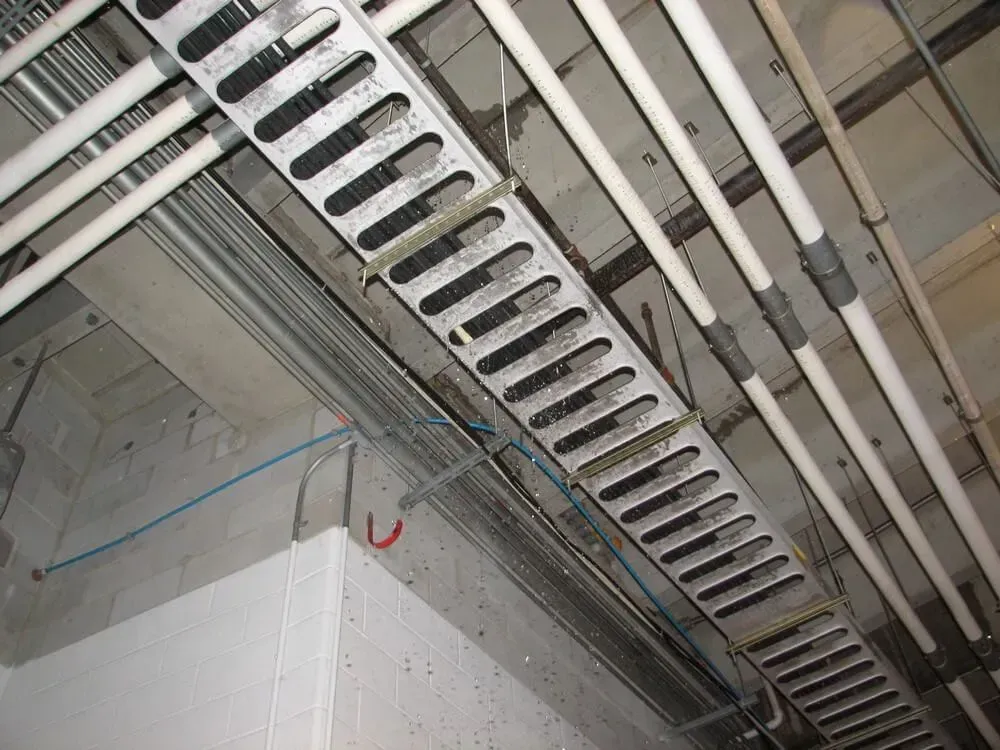 Overhead view of a slotted metal cable tray with conduits and pipes attached to a concrete ceiling.