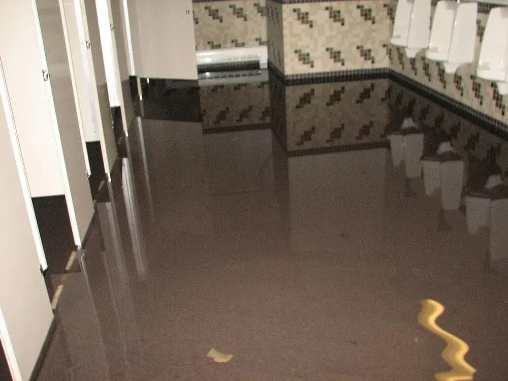 Flooded public restroom with stalls, urinals, and decorative tile. Floor covered in brown water.