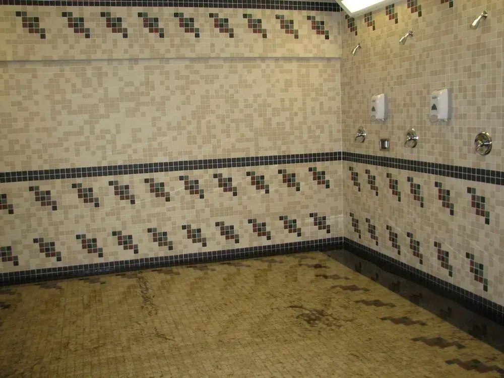 Public shower room with beige tiled walls and floor, multiple showerheads. Decorative dark blue and brown tile borders.