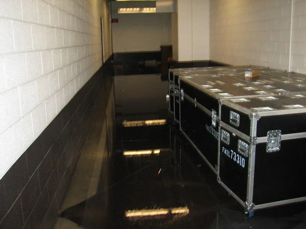 Hallway flooded with dark liquid, large equipment cases sit to the right. White brick wall to the left.