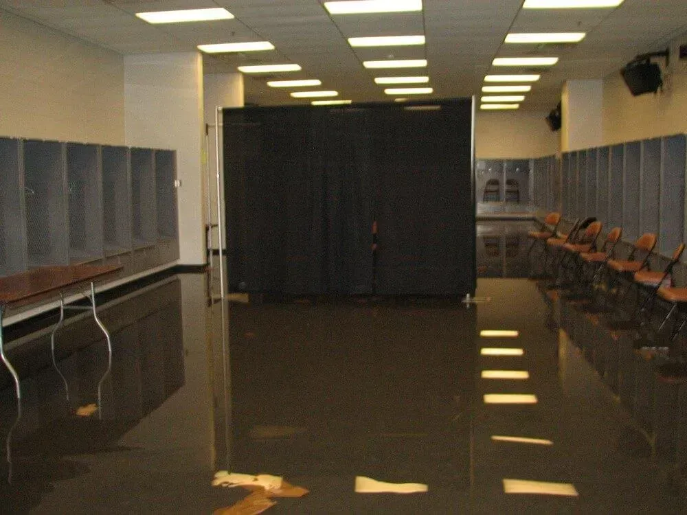 Flooded locker room with rows of lockers, chairs, and a dark curtain. Reflective water covers the floor.