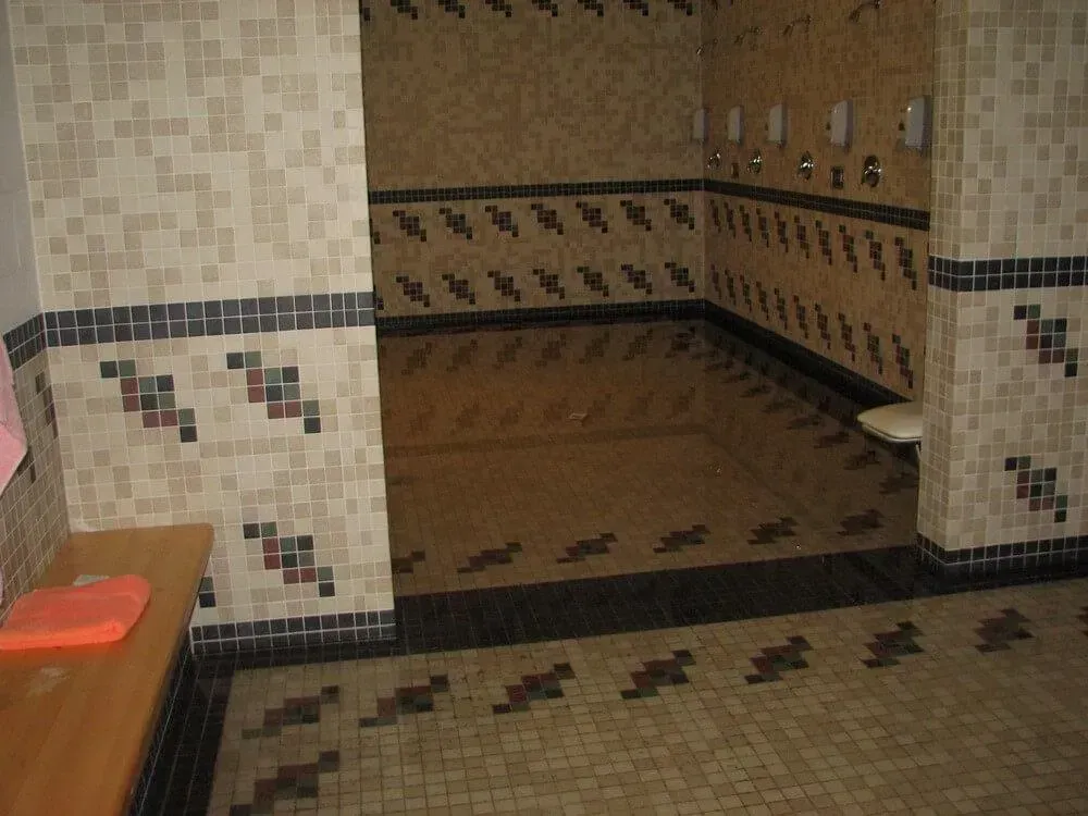 Public shower room with beige and black tiled walls and floor, shower heads, and bench.