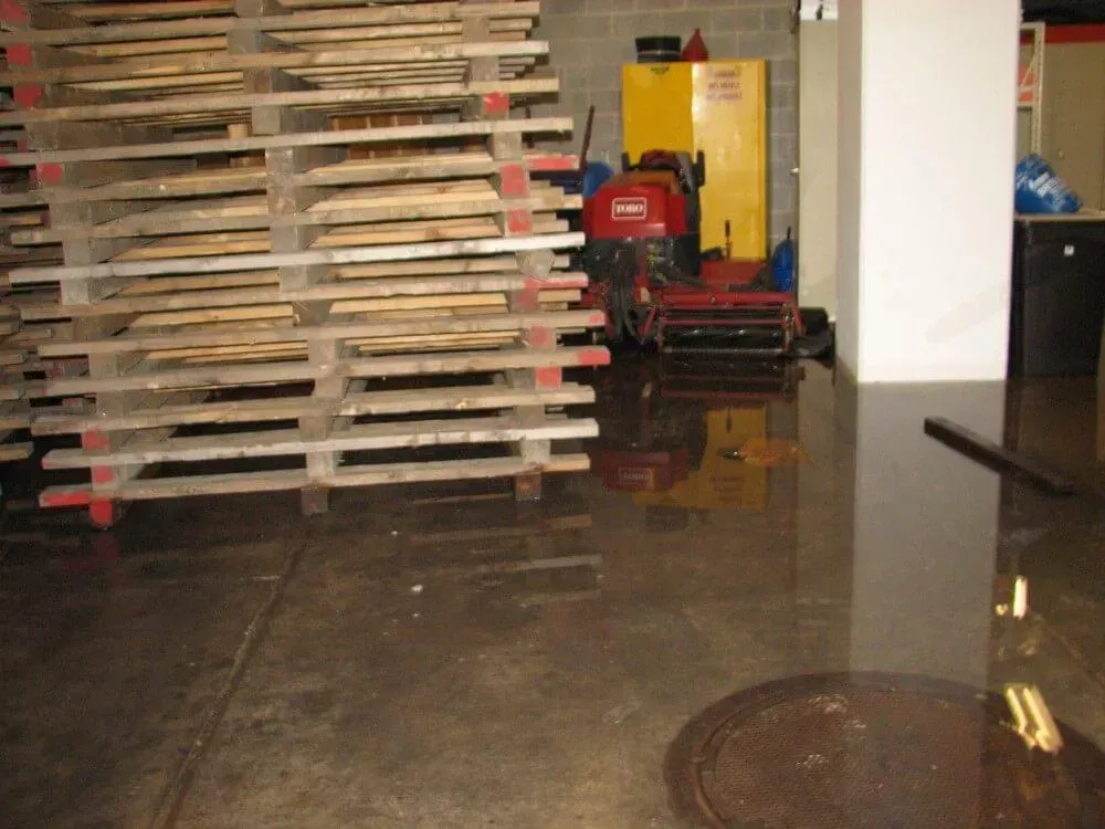 Flooded warehouse floor. Wooden pallets stacked, a red and yellow machine, and a white cabinet reflected in the water.