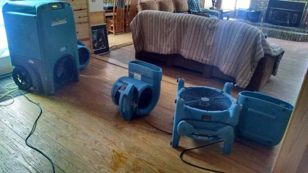 Blue dehumidifiers and fans drying a hardwood floor in a living room, near a couch.