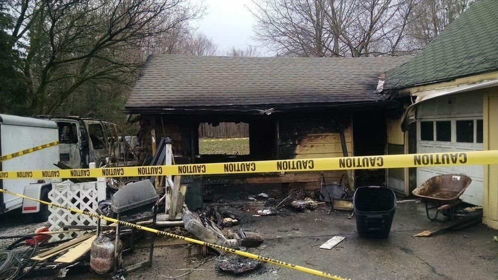 Exterior view of a charred building with caution tape. Debris and burned items are scattered around.