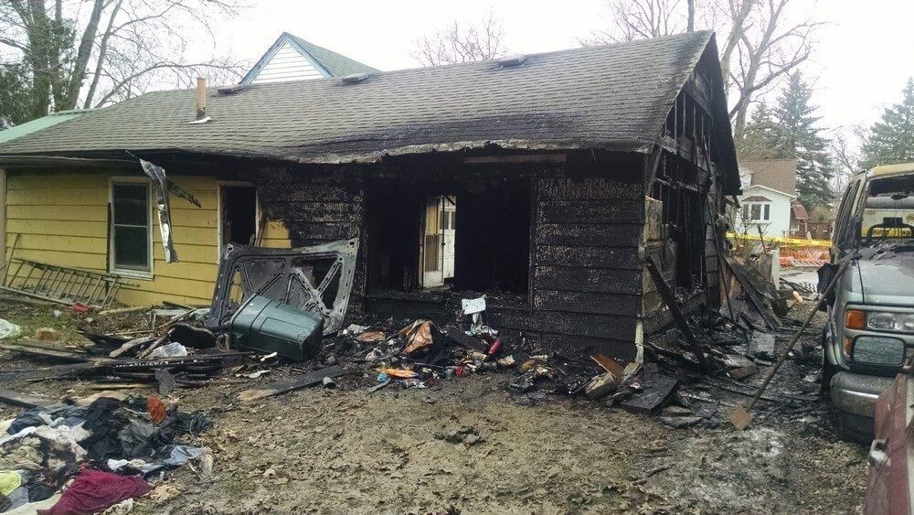 Heavily damaged house after a fire. Charred wood, debris, and burnt belongings are visible.